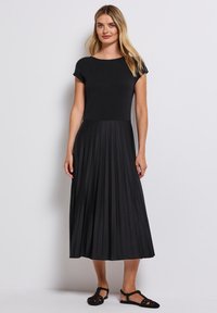 Robe noire avec un haut cintré et une jupe plissée, fabriquée dans un tissu doux. Manches courtes et design jusqu'au genou. Sandales noires décontractées.