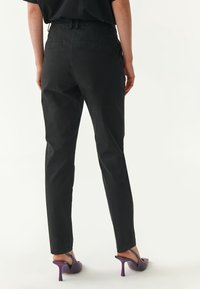 Schwarz maßgeschneiderte Hose mit glatter Textur, ausgestattet mit Gesäßtaschen und schmaler Passform. Kombiniert mit lila High Heels.