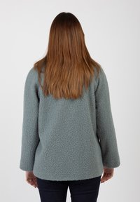 Giacca in felpa di un azzurro chiaro, texture morbida, vestibilità ampia, maniche lunghe, orlo arrotondato, vista da dietro. Jeans di denim visibili sotto.