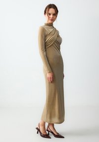 Robe dorée à manches longues avec des détails drapés et une silhouette ajustée, fabriquée dans un tissu texturé et scintillant. Portée avec des escarpins à bout pointu.