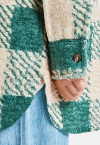 Groen en crèmekleurig geruit fleecejack met een textuurstof, knoopmanchetten en een gebogen zoomlijn. Close-up van het mouwdetail met een hand.