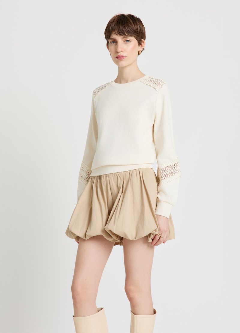 Sweat-shirt crème avec détails en dentelle sur les épaules, associé à une jupe ballon beige. La tenue est dotée d'une ceinture cintrée et d'un ourlet volumineux.