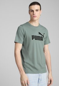 Vihreä puuvillapaita, jossa on musta PUMA-logo edessä. Lyhyet hihat ja pyöreä kaula-aukko. Mallilla on päällä vaaleat farkut.