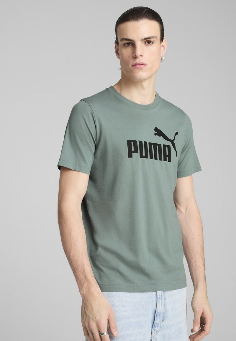 Vihreä puuvillapaita, jossa on musta PUMA-logo edessä. Lyhyet hihat ja pyöreä kaula-aukko. Mallilla on päällä vaaleat farkut.