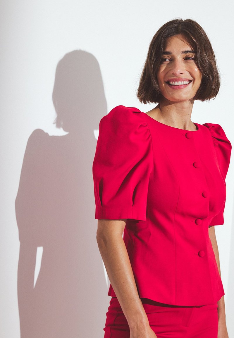 Blusa rossa a maniche a sbuffo con silhouette aderente, caratterizzata da bottoni rotondi e una texture liscia, che proietta un'ombra delicata su uno sfondo bianco.