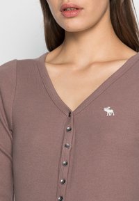 Frau, die ein geripptes, mauvefarbenes V-Ausschnittoberteil mit einer Reihe von Knöpfen und einem kleinen, weißen, gestickten Elch-Logo auf der linken Brust trägt.