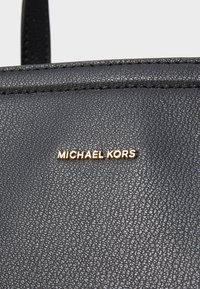 Schwarze, strukturierte Lederhandtasche mit einem goldenen Logo-Detail. Eine Nahaufnahme zeigt die Nähte und die griffige Oberflächenstruktur.