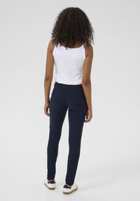 Marineblaue, enganliegende Hose aus dehnbarem Material mit glatter Textur und elegantem Design, kombiniert mit einem weißen Tanktop und lässigem Schuhwerk.