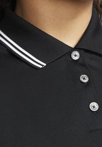 Černé polo tričko s žebrovaným límečkem, který má dva bílé pruhy a tři viditelné knoflíkové zapínání na légě.