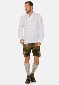 Weißes Leinenhemd mit langen Ärmeln und Knopfleiste, braune Lederbermudas mit dekorativer Stickerei und graue gestrickte Socken. Braune Schuhe runden das Outfit ab.