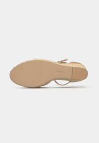 Sandal avec semelle en caoutchouc beige, talon compensé en espadrille, et fermeture à boucle de bride de cheville, vu par le dessous.