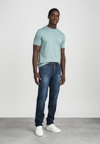 Svijetloplava majica kratkih rukava s malim logotipom, u paru s tamnoplavim trapericama i bijelim tenisicama na neutralnoj pozadini.