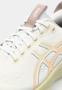 White running shoe featuring a textured mesh upper, gray and yellow accents, lace closure, and a pink pull tab.
