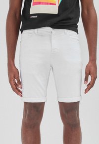 Pantalones cortos de mezclilla blanca con puños enrollados, diseño de cinco bolsillos, cierre con botón metálico y cremallera. Llevado con una camiseta gráfica negra.