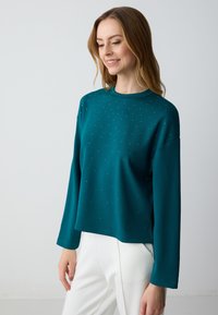 Felpa lunga colore teal con scollo rotondo aderente, decorata con accenti di strass sparsi e una vestibilità comoda. Indossata con pantaloni bianchi.