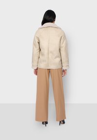 Giacca in finta pelliccia shearling beige con ampio colletto, foderata in crema e con polsini. Il modello indossa pantaloni beige a gamba larga e tacchi alti.