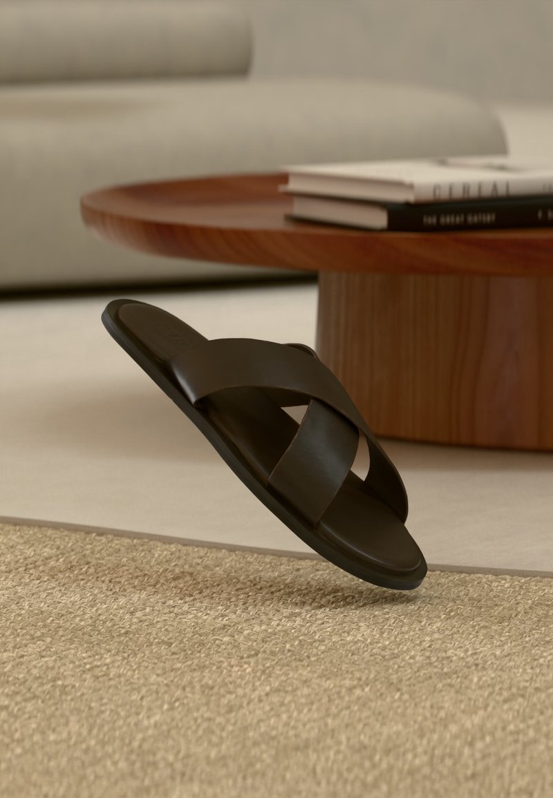 Μαύρο δερμάτινο σανδάλι slide με φαρδιές σταυρωτές λουρίδες σε μπεζ χαλί, κοντά σε στρογγυλό ξύλινο τραπεζάκι σαλονιού με στοιβαγμένα βιβλία.