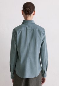 Hellblaues Hemd mit Kragen aus weichem Stoff, langärmlig und mit abgerundetem Saum; die Rückansicht zeigt Knopfdetails und eine glatte Textur.