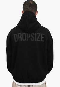 Sort hættejakke med stort "DROPSIZE" tekst på ryggen. Løs pasform, snor i hætten og ribbet kant. Blødt materiale og tekstureret stof.