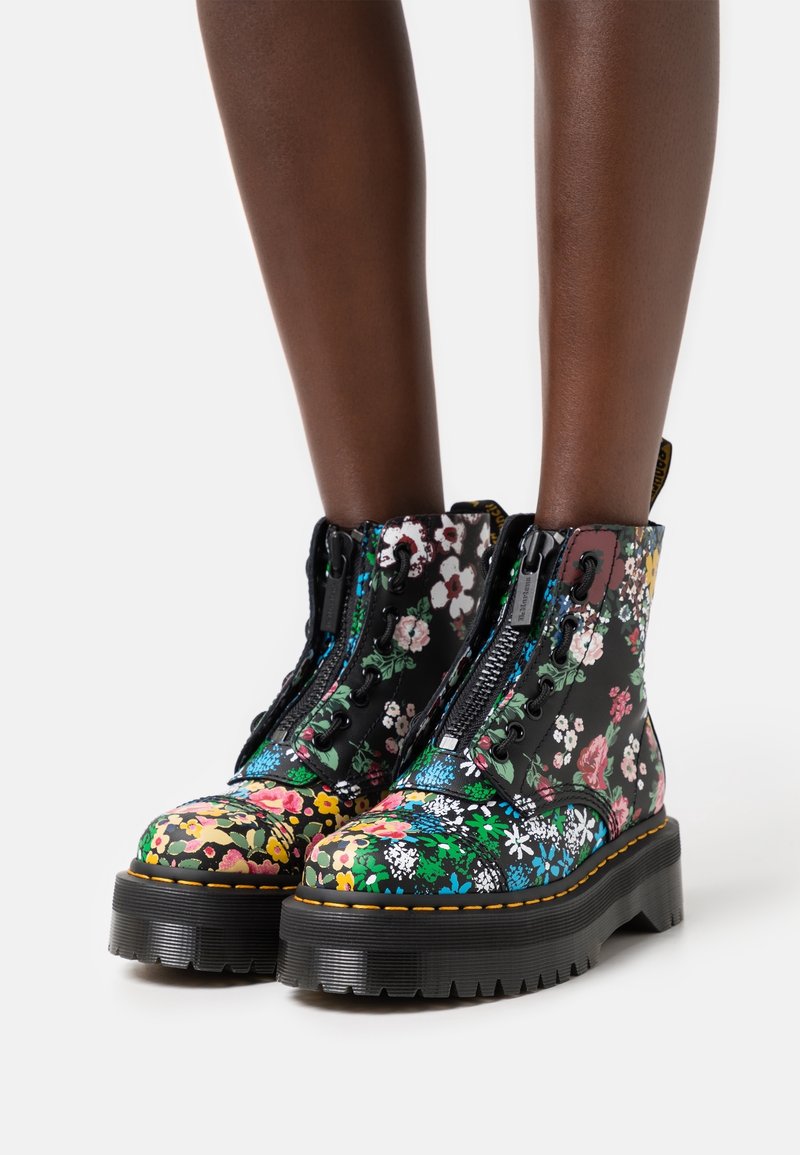 Bottes à plateforme noires avec imprimé floral multicolore, fermetures éclair à l'avant, semelles épaisses, portées sur des jambes à la peau foncée.