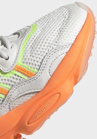 Harmaa lenkkari, jossa verkkomateriaalia päällisosassa, neonvihreät ja oranssit yksityiskohdat, teksturoitu kumipohja ja näkyvä brändäys kantapäässä.