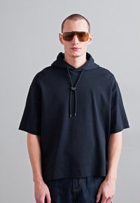 Mustkas suuruses kapuutsiga sviiter lühikeste varrukatega, stringiga kapuuts ja tekstuuriga kangas. Kanda pruunikate prillidega.