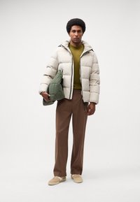 Puffer jacket in light gray with a zipper, paired with a green quilted bag. Features a hood and side pockets. Brown trousers and beige shoes.