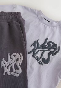 Voltitud halli vahvlitehnikas pükse ja helelilla t-särk, mõlemal kangal reljeefsed mustad ja valged "ALSANITS LONDON" logod.