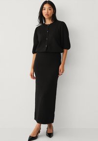 Schwarzes Zweiteiler-Outfit, bestehend aus einem strukturierten Kurzarmoberteil mit Knopfverschluss und einem enganliegenden Maxikleid; kombiniert mit spitz zulaufenden schwarzen Absatzschuhen.