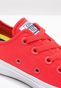 Red canvas sneaker with a low-top design, featuring rubber eyelets and a white rubber sole. Includes a label with a blue graphic.
