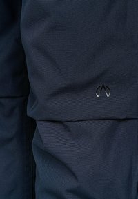 Navy stof tæt på med en tekstureret overflade, og et lille sølvemblem, der viser et stiliseret logo nederst til højre.