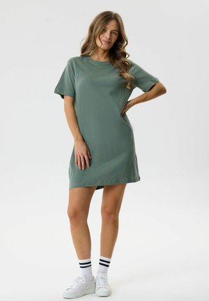 Femme debout, main sur la hanche, portant une robe T-shirt verte à manches courtes, baskets blanches et chaussettes blanches à rayures noires sur fond uni.