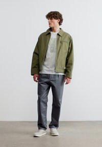 Άνδρας με σγουρά μαλλιά που φοράει πράσινο μπουφάν με φερμουάρ, λευκό μπλουζάκι με σχέδιο, γκρίζο παντελόνι και λευκά αθλητικά παπούτσια, στέκεται σε γκρίζο δάπεδο.