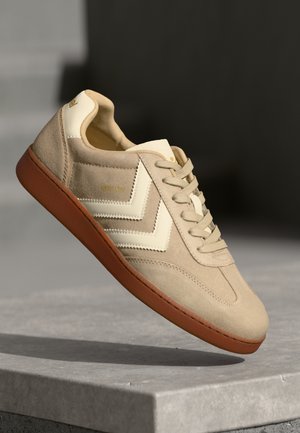 Beige suede sneaker with cream leather accents, brown rubber sole, rounded toe, and tonal stitching. Features a chevron design on the sides.