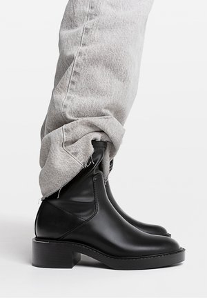 Black leather ankle boots worn with light gray rolled-up jeans against plain white background.