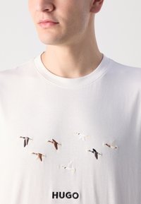 Balts kokvilnas T-krekls ar izšūtiem putniem brūnā, pelēkā un baltā krāsā virs melna "HUGO" logo apakšā centrā.