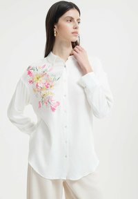 Weißes Hemd mit Knöpfen aus leichtem Stoff, mit rosa und gelben Blumenstickereien auf der linken Schulter. Abgerundeter Saum.