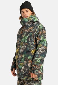 Mężczyzna ubrany w zimową kurtkę i spodnie w wzór kamuflażu z motywem szyszek i liści, czarną czapkę oraz rękawiczki bezpalcowe.