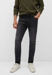 Muž v tmavě šedých slim-fit džínech, olivově zeleném svetru a bílých teniskách, s jednou rukou v kapse.