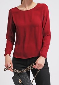 Rote Seidenbluse mit langen Ärmeln, rundem Ausschnitt und glatter Textur; kombiniert mit einer schwarzen, gesteppten Handtasche mit einer Kettenriemen.