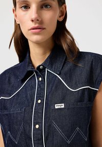 Dyp denim ermeløs skjorte med hvit piping, spiss krage, to brystlommer og en liten Wrangler-logo på venstre lomme.