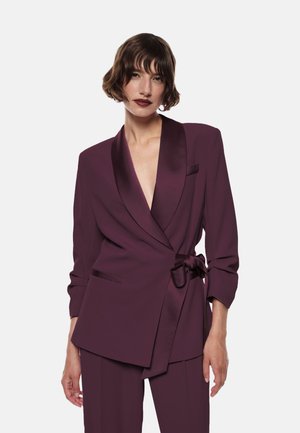 Donna con i capelli corti castani che indossa un giacca di un profondo viola con rever in satin e una cintura annodata in vita, abbinata a pantaloni coordinati.