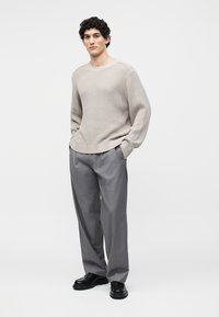 Homme debout portant un pull en maille beige, un pantalon gris ample, des chaussures noires, les mains dans les poches, expression neutre, fond blanc uni.