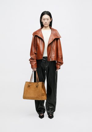 Giacca di pelle marrone con colletto oversize, abbinata a un maglione chiaro e pantaloni di pelle nera. Tieni in mano una borsa in suede beige con manici marroni.