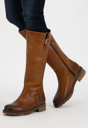 Høje brune læderstøvler med sidelås og spænde, båret med mørkeblå jeans mod hvid baggrund.