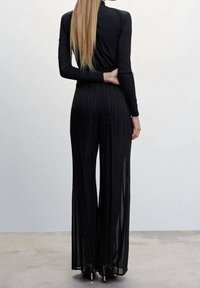 Femme aux longs cheveux blonds portant un haut à manches longues noir et un pantalon noir large à taille haute, se tenant devant un fond uni.