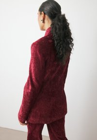 Raudono aksomo kostiumas su tekstūruotu paviršiumi, apimantis pritaikytą švarką ir atitinkančias kelnes. Dizainas yra elegantiškas, su aštriais apykaklėmis ir prigludusia siluetu.