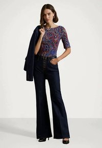 Žena v pestrých vzorovaných topoch, s tmavými vysokými rozšírenými džínami, čiernymi lodičkami, drží modrý blejzer cez jedno plece.