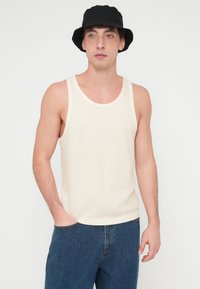 Μπεζ αμάνικη μπλούζα από ριμπ ύφασμα με στρογγυλή λαιμόκοψη, σε συνδυασμό με σκούρο μπλε τζιν και μαύρο καπέλο τύπου bucket.