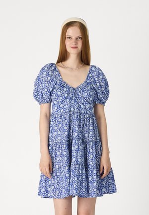 Sininen kukalline mekko puffihihan kanssa, vyötäröltä napakka, ja kerroksellinen helma. Omistaa neliön muotoisen kaula-aukon ja monimutkaisia valkoisia kuvioita.